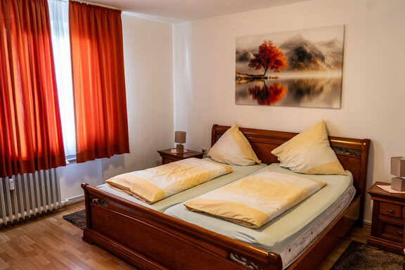 Doppelzimmer mit großem Holzbett, Nachttischen, roten Vorhängen und Wandbild.